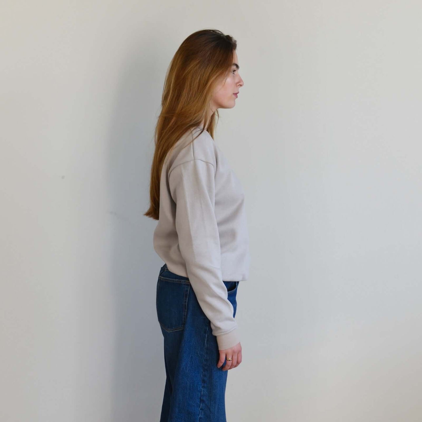 Woman wearing a light gray sweater and blue jeans against a plain white wall