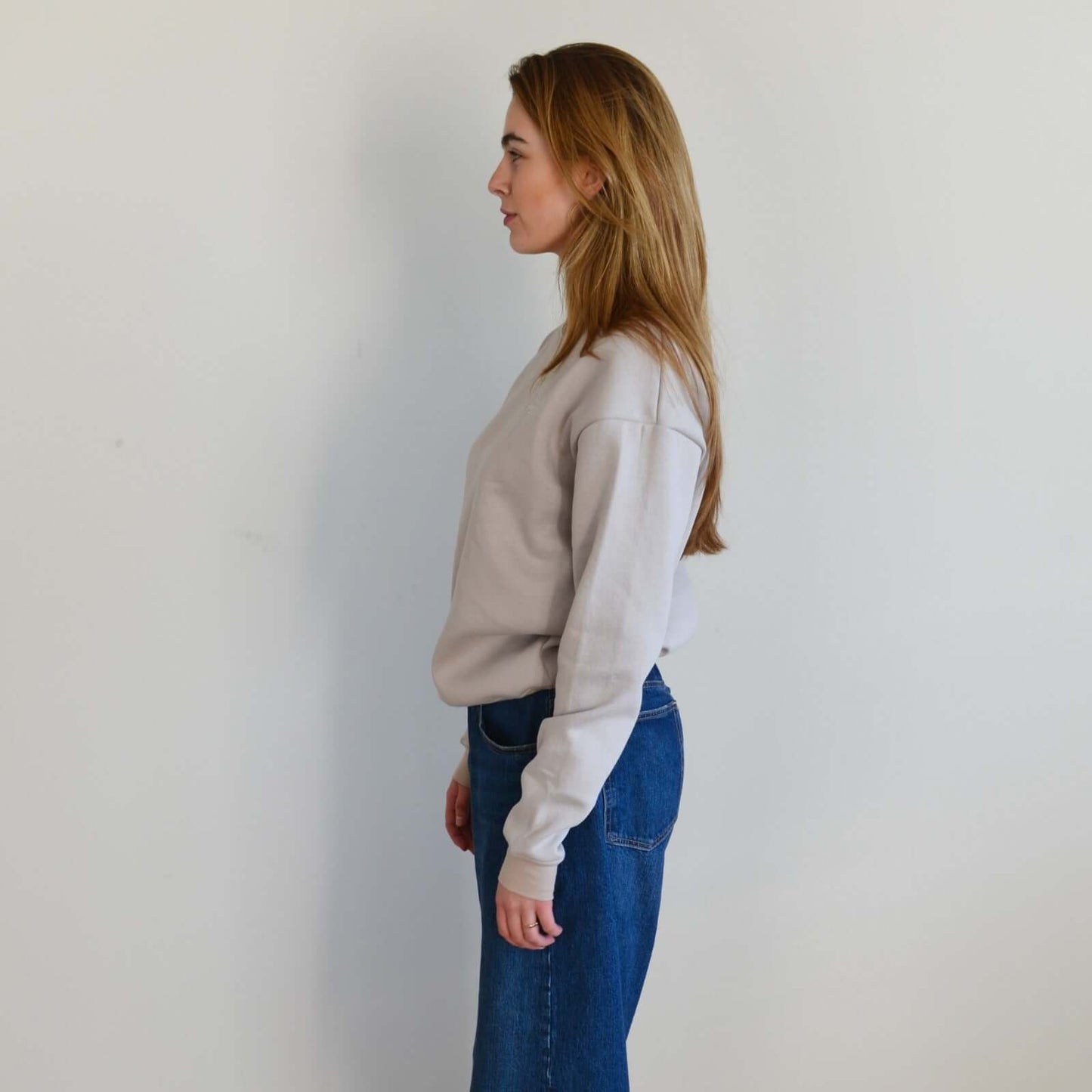 Woman wearing a beige sweater and blue jeans against a plain white wall