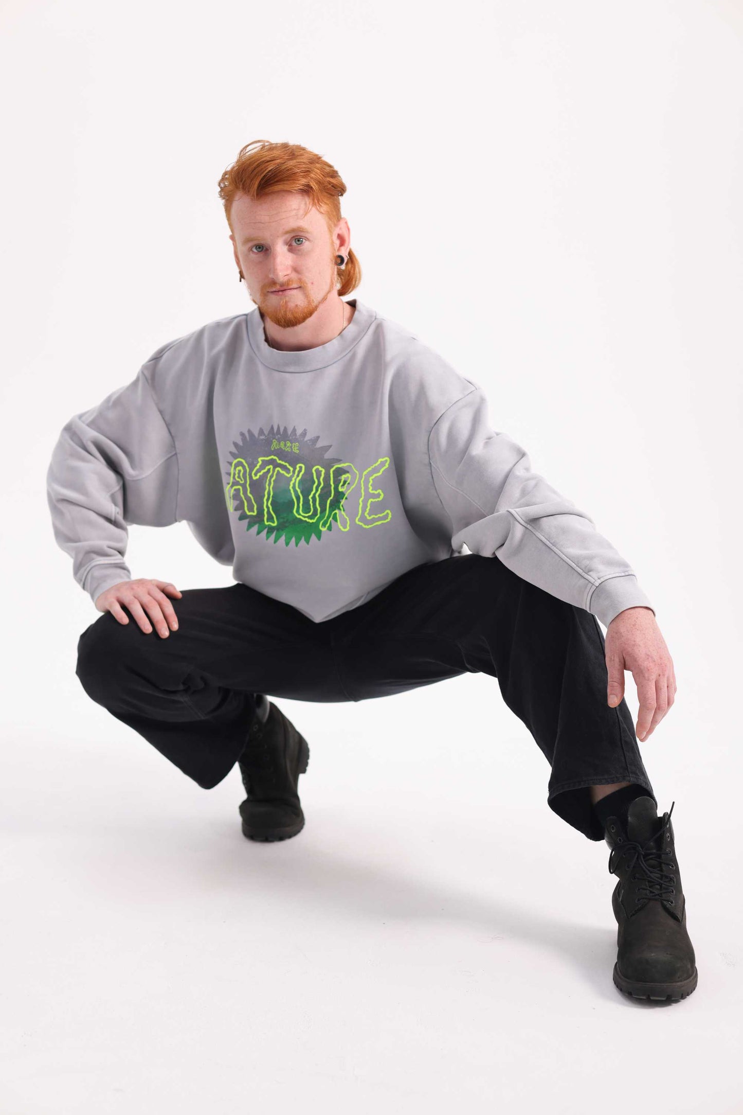Man wearing a gray sweatshirt with green text, black pants, and black boots on a white background
