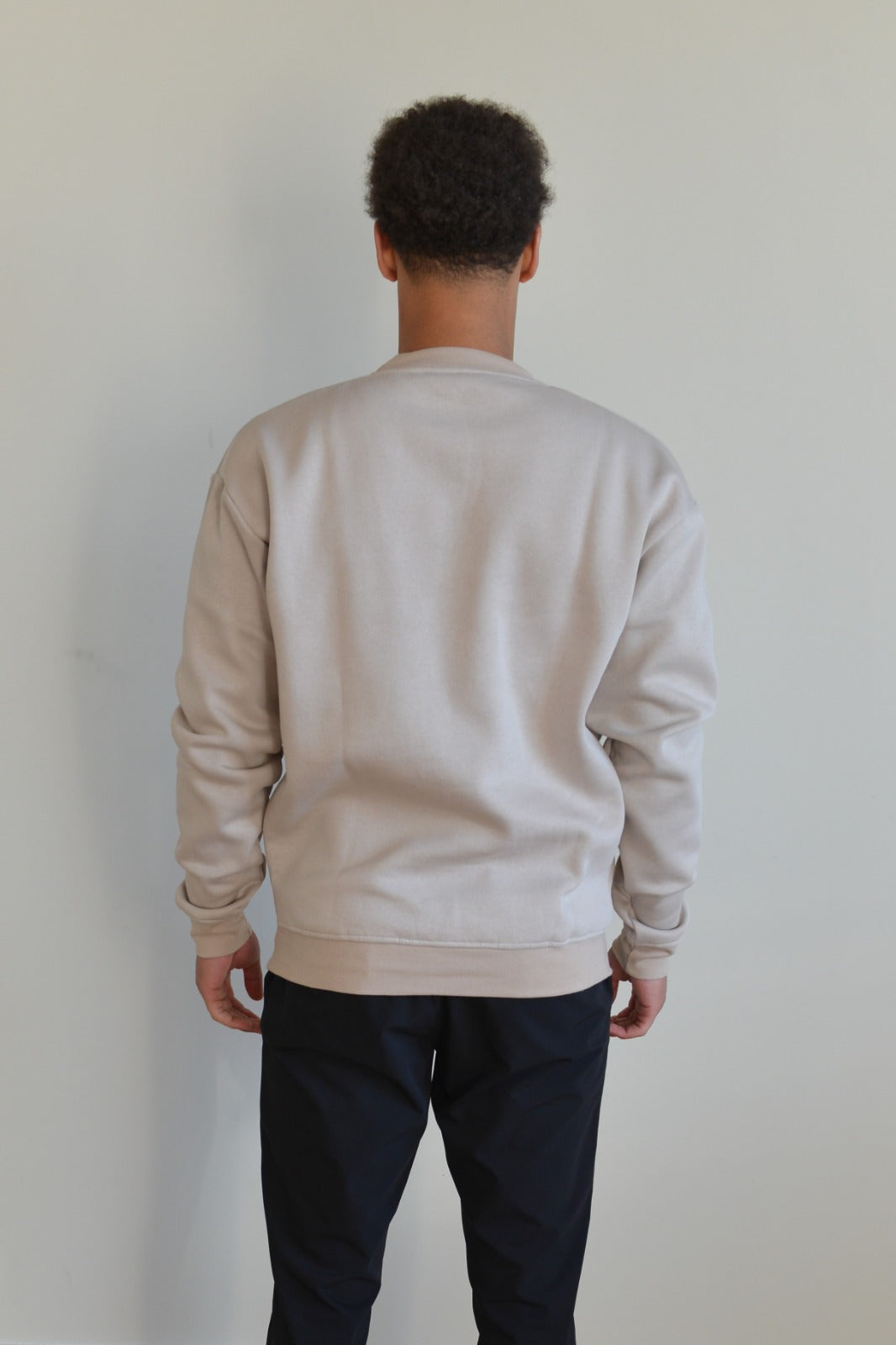 Person wearing a beige sweatshirt and dark pants against a plain background