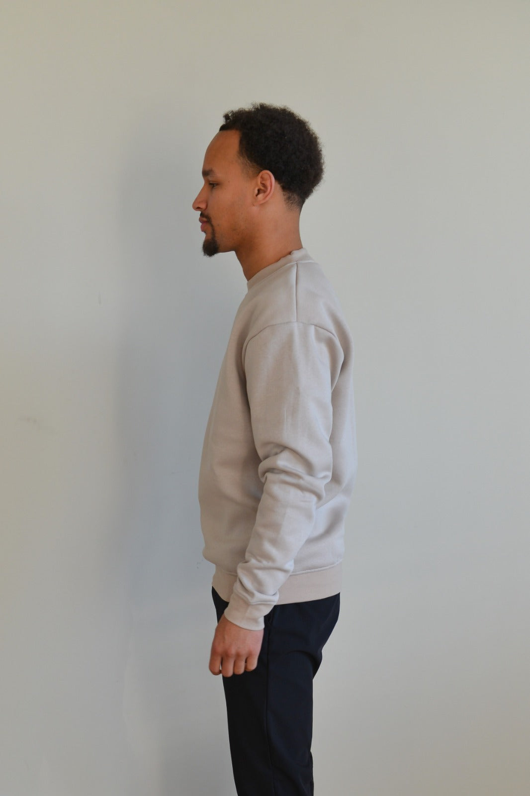 Man wearing a light gray sweatshirt and dark pants against a plain white wall