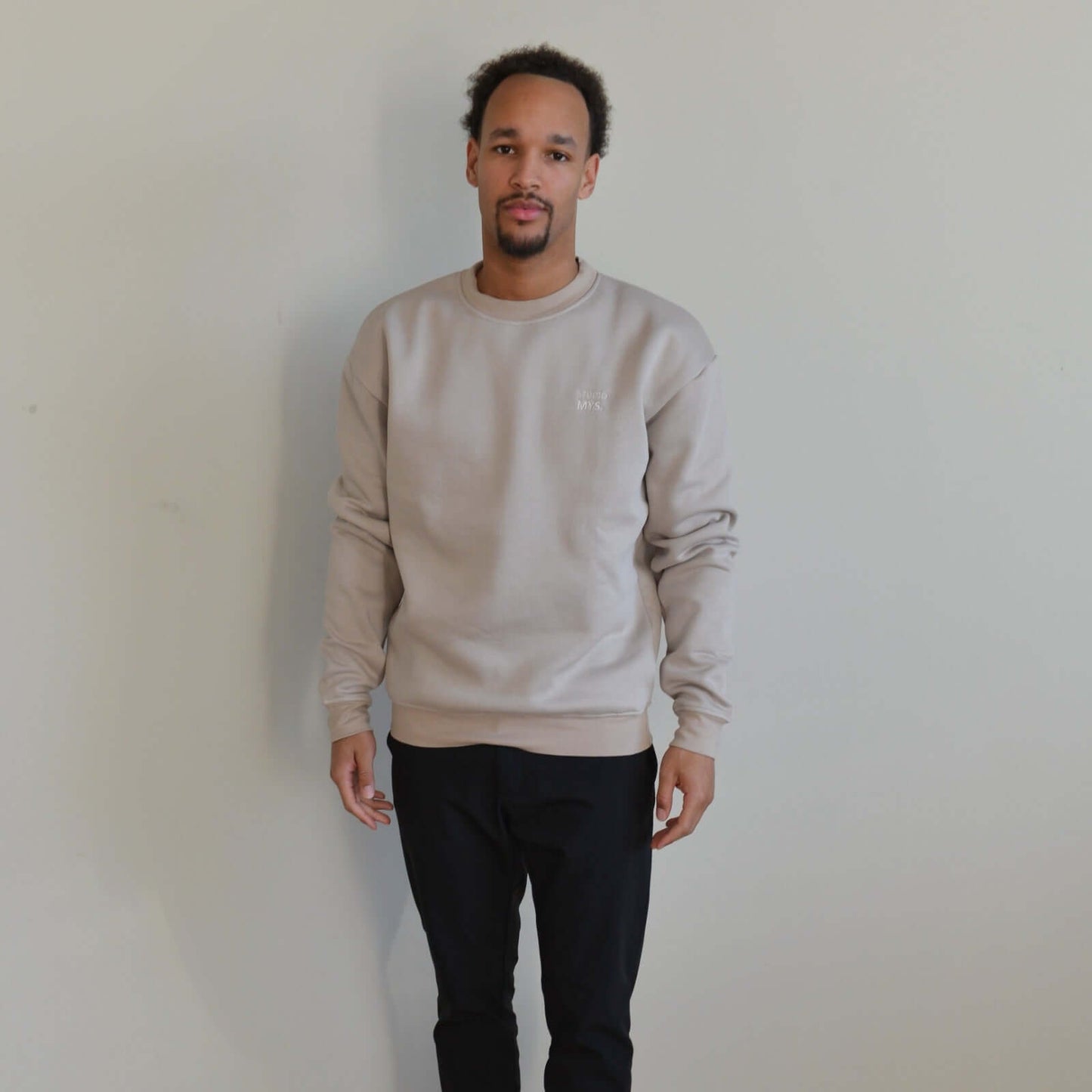 Man wearing a beige sweatshirt and black pants against a plain background