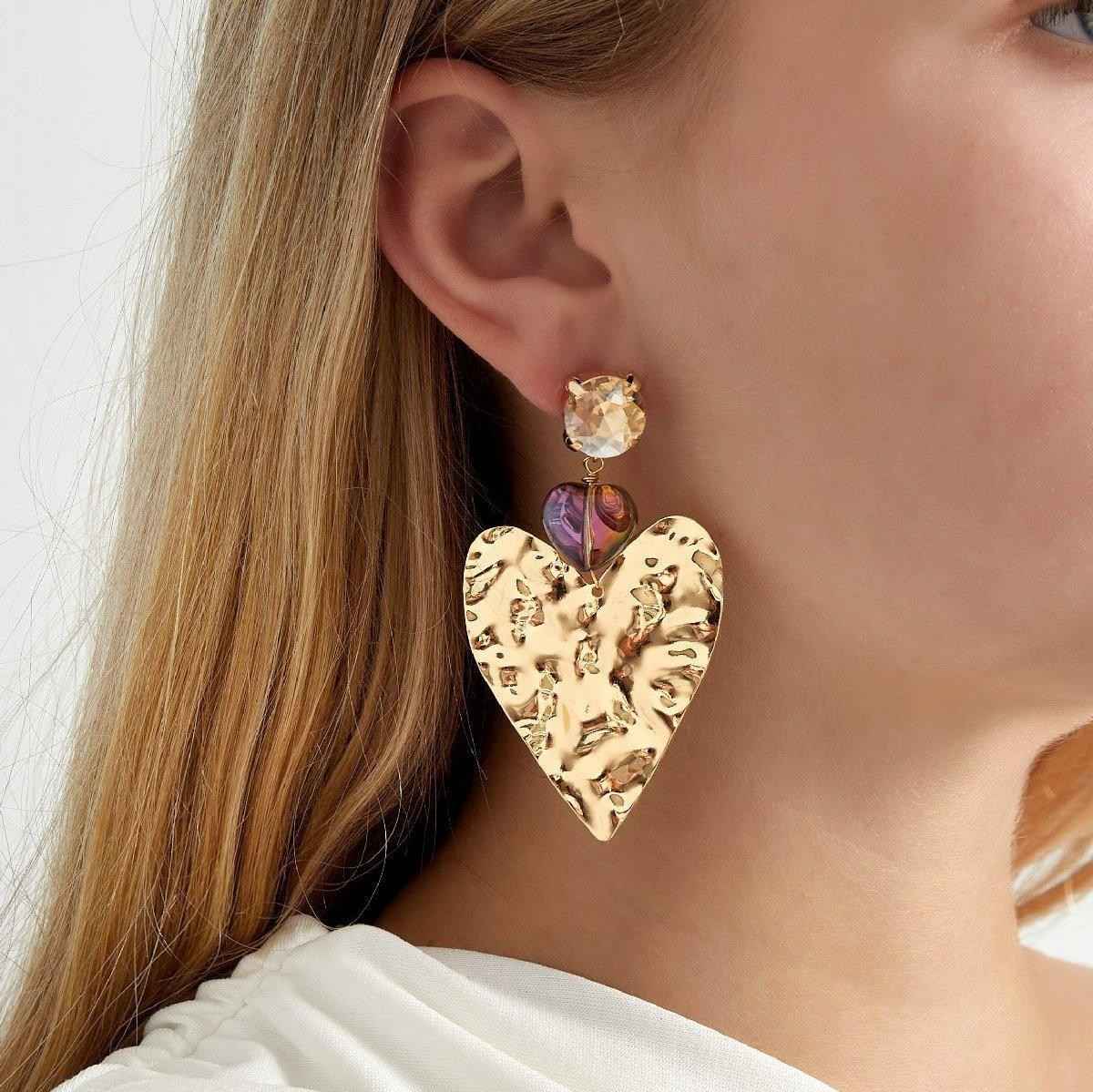 Gold heart-shaped earrings with colorful gemstones worn by a model.