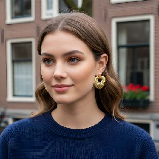 Woman wearing gold heart earrings in front of a building with flowers.