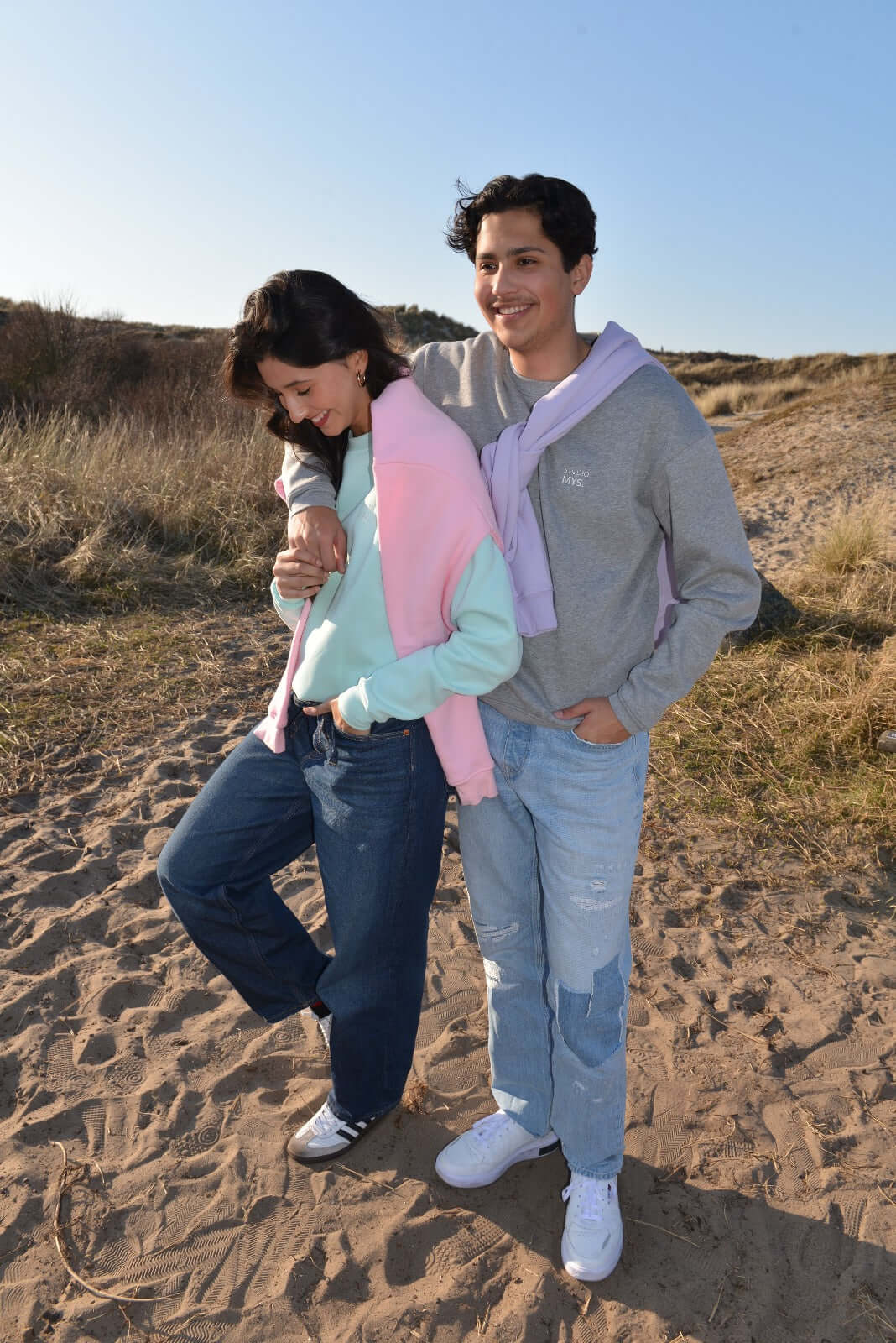 Two people standing on a sandy beach with a clear blue sky wearing sweatshirts