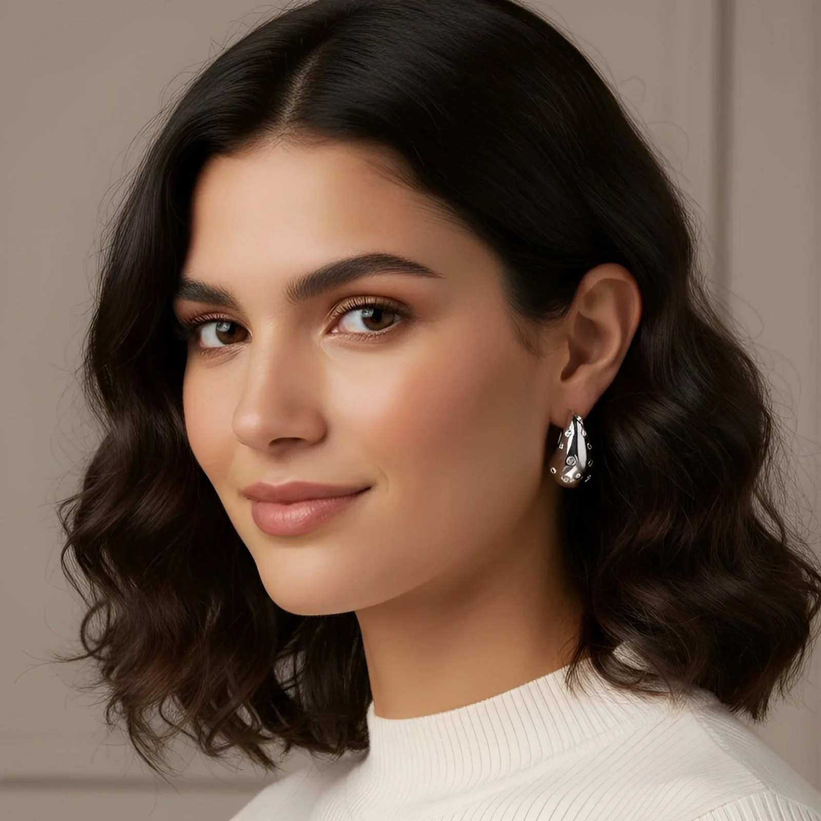 Woman with wavy hair wearing a white sweater and silver earrings against a neutral background